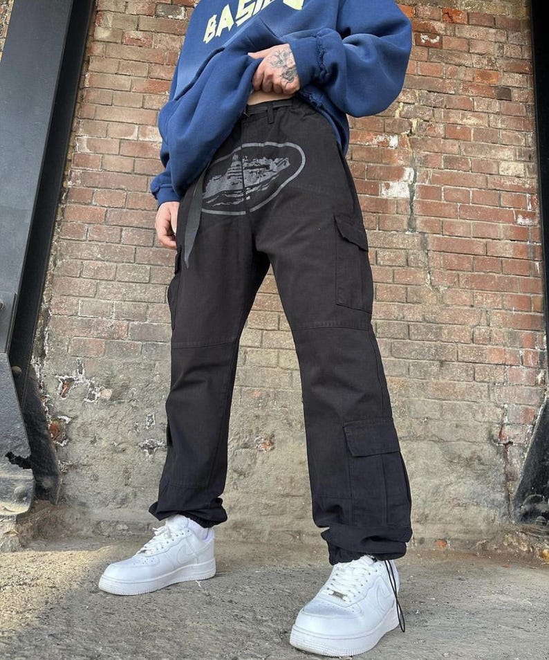 Puede incluir: Pantalones cargo negros con un dise&ntilde;o gr&aacute;fico gris en la parte delantera, con m&uacute;ltiples bolsillos y un cintur&oacute;n. Los pantalones se combinan con zapatillas blancas y una sudadera azul. El fondo es una pared de ladrillos.