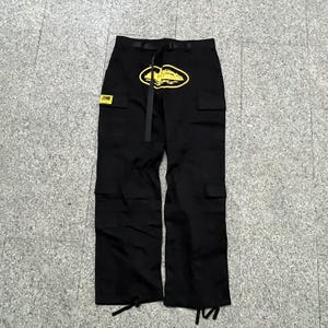 Puede incluir: Pantalones cargo negros con un logotipo amarillo en la parte delantera y un parche a juego. Los pantalones tienen m&uacute;ltiples bolsillos, un cintur&oacute;n y cordones en los tobillos. El logotipo tiene forma ovalada con un dise&ntilde;o en el interior.