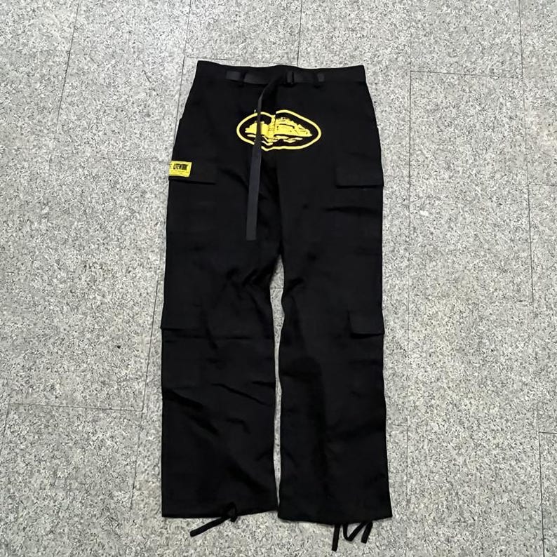 Puede incluir: Pantalones cargo negros con un logotipo amarillo en la parte delantera y un parche a juego. Los pantalones tienen m&uacute;ltiples bolsillos, un cintur&oacute;n y cordones en los tobillos. El logotipo tiene forma ovalada con un dise&ntilde;o en el interior.