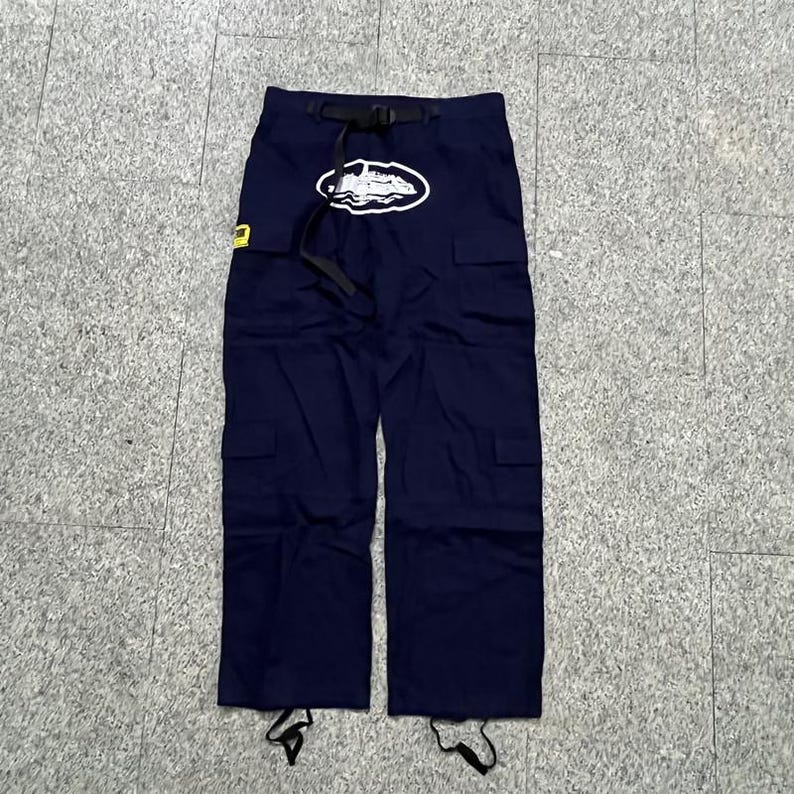 Puede incluir: Pantalones cargo azul marino con cintur&oacute;n negro y logotipo blanco. Los pantalones tienen m&uacute;ltiples bolsillos y cordones en los tobillos. Se ve un parche amarillo en el lado izquierdo.