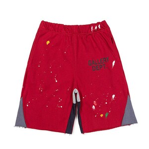 May include: Red athletic shorts with paint splatter details and the text "GALLERY DEPT." in black. The shorts have grey and navy blue side panels and a drawstring waistband. A casual style.