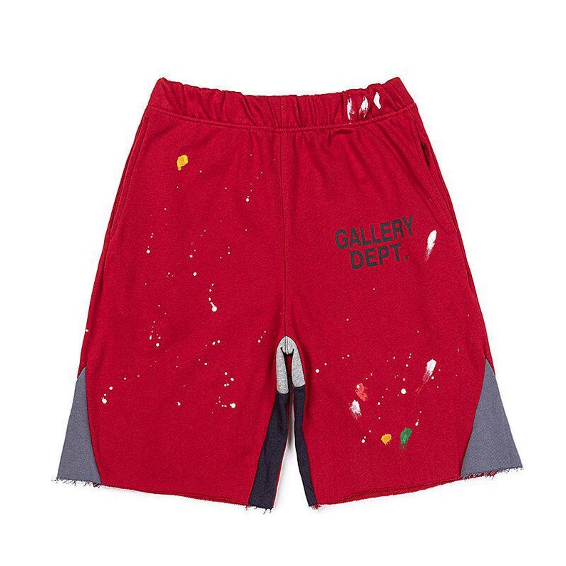 May include: Red athletic shorts with paint splatter details and the text "GALLERY DEPT." in black. The shorts have grey and navy blue side panels and a drawstring waistband. A casual style.