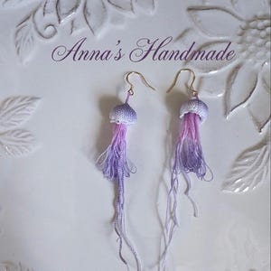 May include: Handmade jellyfish earrings. Each earring has a small, crocheted, lavender and white cap with long, flowing, ombre purple and white strands. The earrings hang from gold-coloured ear wires. The text "Anna's Handmade" is visible.