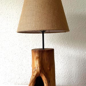 May include: A rustic table lamp featuring a natural wood base and a burlap lampshade. The base is a carved tree stump with a dark opening. The lampshade is a light brown color. The lamp is designed for indoor use.