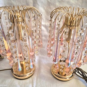 May include: A pair of table lamps featuring gold-toned metal frames with cascading clear and iridescent pink glass teardrop prisms and beaded strands. Each lamp has a circular gold base and a curved wire top structure holding the hanging glass elements.