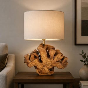 Op de afbeelding: Een tafellamp met een cilindervormige beige stoffen kap en een voet gemaakt van een natuurlijk, ruw stuk lichtbruin hout met zichtbare nerven en organische, onregelmatige randen, ondersteund door een dunne, messingkleurige metalen stang.