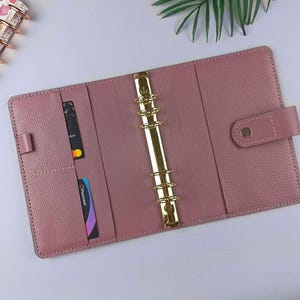 Può includere: Un raccoglitore A6 color rosa polvere con finitura testurizzata, dotato di un meccanismo metallico dorato a sei anelli al centro. L'interno include diverse fessure verticali per carte sul lato sinistro e una chiusura a bottone a pressione a destra.
