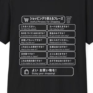 Könnte beinhalten: Ein schwarzes T-Shirt mit kurzen Ärmeln und einem weißen Grafikdruck, der einen Einkaufs-Sprachführer zeigt. Das Design enthält eine Liste nützlicher japanischer Sätze mit englischen Übersetzungen, angeordnet in einem Raster mit Symbolen.
