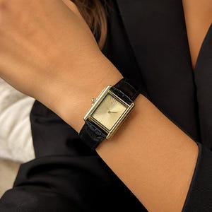 Può includere: Un orologio da polso rettangolare dorato con cinturino in pelle nera. Il quadrante dell'orologio presenta un motivo a raggiera. L'orologio è su un polso, con un blazer nero sullo sfondo. Un accessorio classico ed elegante.