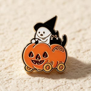 May include: A gold-toned metal enamel pin featuring a white ghost wearing a black witch hat and a small black cat sitting together inside a bright orange jack-o'-lantern pumpkin that is mounted on four small wheels, creating a rolling cart design.