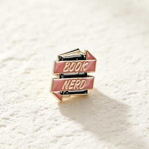 May include: A gold-toned metal enamel pin shaped like a stack of black books wrapped in two pink ribbons. The top ribbon features the word BOOK in gold lettering, and the bottom ribbon features the word NERD in gold lettering, with a gold border.