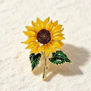 Op de afbeelding: Een zonnebloem broche met felgele bloemblaadjes, een donkerbruin textuurcentrum en twee groene bladeren aan een goudkleurige steel. Het ontwerp is uitgevoerd in emaille met goudkleurige metalen omlijningen en toont de bloem in een realistische vorm.