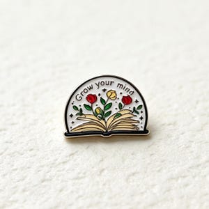 May include: A semi-circular enamel pin featuring an open book with yellow pages and green stems sprouting red and yellow roses. The white background includes small black stars and the text "Grow your mind" arched along the top edge.
