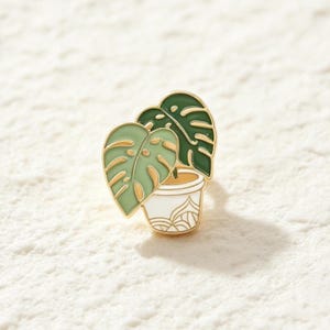 May include: A metal enamel pin featuring a potted monstera plant. The plant has two leaves, one in light green and one in dark green, with gold-toned outlines. The pot is white with gold-toned decorative line details at the base and rim.