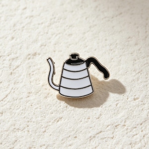 May include: A white and black enamel pin shaped like a gooseneck kettle. The kettle features a white body with horizontal black stripes, a black lid, and a black handle. The design is outlined in a thin metallic border, creating a clean, graphic look.