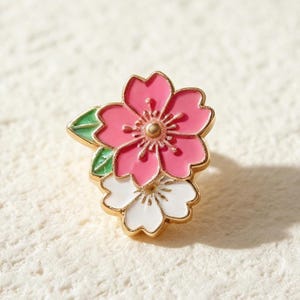 Puede incluir: Un broche de esmalte y metal dorado que presenta una flor de cerezo rosa superpuesta sobre una flor de cerezo blanca más pequeña, acentuada con dos hojas verdes. El diseño tiene un contorno de metal dorado y un estambre central dorado.