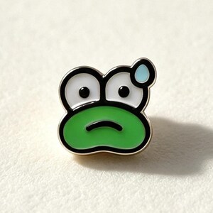 Könnte beinhalten: Ein Emaille-Anstecker aus Metall in Form eines grünen Froschgesichts mit großen weißen Augen, schwarzen Pupillen und einem kleinen blauen Schweißtropfen über dem linken Auge. Der Frosch hat einen geraden, schwarzen Mundstrich.