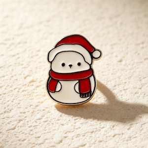 May include: A metal enamel pin shaped like a white bear wearing a red and white Santa hat and a red scarf. The bear has simple black dot eyes and a small black nose. The pin features gold-toned metal outlines around the white, red, and black enamel.