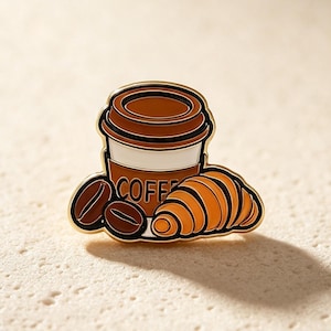 Pode incluir: Um broche de esmalte e metal dourado com uma chávena de café castanha e branca com o texto COFFEE impresso na lateral, acompanhado por dois grãos de café castanho escuro e um croissant dourado, tudo contornado por um rebordo de metal preto.