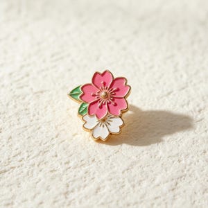 May include: A gold-toned metal enamel pin featuring two layered cherry blossom flowers, one in vibrant pink and one in white, accented with a small green leaf. The design showcases detailed petals and a central stamen, creating a floral accessory.