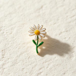 May include: A gold-toned metal enamel pin shaped like a daisy flower. The pin features white petals, a bright yellow center, and a green stem with two small leaves. The design is outlined in thin gold metal, creating a clean and defined appearance.