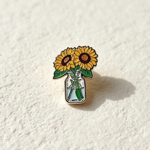 Op de afbeelding: Een metalen emaille speld met twee gele zonnebloemen met bruine harten en groene bladeren, geplaatst in een doorzichtige glazen pot gevuld met water. Het ontwerp is omlijnd met goudkleurig metaal en toont de stelen door het glas heen.
