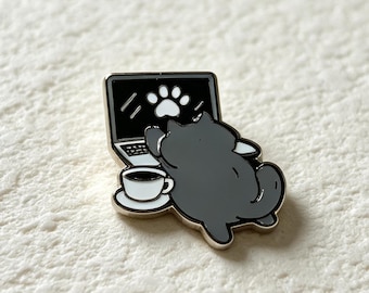 May include: A metal enamel pin featuring a dark gray cat lying down in front of an open laptop. The laptop screen displays a white paw print icon. A small white coffee cup on a saucer sits next to the laptop. The pin has a metallic silver outline.
