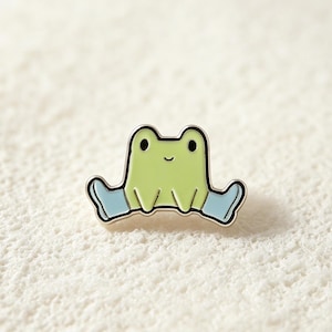 May include: A charming frog pin featuring a light green body, black eyes, and a smiling mouth. The frog is wearing light blue boots. The pin has a silver outline and is set against a white textured background.