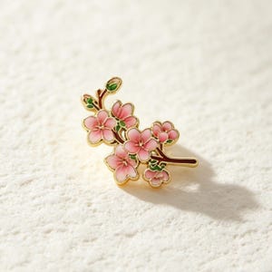 Puede incluir: Un broche de esmalte y metal dorado con forma de rama de cerezo en flor. El diseño presenta flores rosas de cinco pétalos con sombreado degradado, pequeños brotes verdes y ramas de color marrón oscuro con contornos dorados en la pieza.