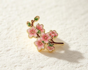 Könnte beinhalten: Eine goldfarbene Emaille-Anstecknadel in Form eines blühenden Kirschblütenzweigs. Das Design zeigt rosafarbene Blüten mit fünf Blütenblättern und Farbverlauf, kleine grüne Knospen sowie dunkelbraune Zweige mit goldenen Umrandungen am gesamten Stück.