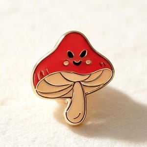 May include: A gold-toned metal enamel pin shaped like a mushroom with a bright red cap and a light beige stem. The mushroom cap features a smiling face with two black eyes and a curved mouth, accented by two small light-colored circular cheeks.