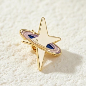 以下が含まれることがあります： 中央にリングを配した四芒星の形をしたゴールドトーンの金属製ピン。星にはライトパープル、ダークブルー、ホワイトのエナメル装飾が施されており、磨き上げられたメタリックな仕上げの天体モチーフのデザインとなっています。