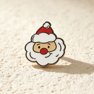 May include: A metal enamel pin shaped like a cartoon Santa Claus head. The design features a red hat with a white pom-pom and trim, a tan face, a bright red nose, and a thick white beard. The entire figure is outlined in gold-toned metal.