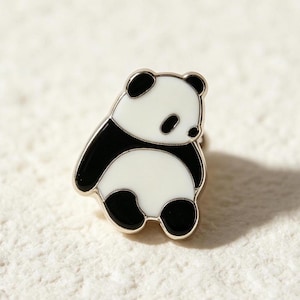 May include: A small enamel pin shaped like a sitting panda bear. The design features a white head and belly with black ears, limbs, and eye patches, all outlined in a thin gold-toned metal border. The panda is depicted in a rounded, seated pose.