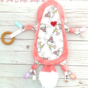 May include: A pink and white baby toy with a soft, fuzzy texture. The toy has a variety of textures and colours, including a wooden ring, ribbons, and a plush tail. The toy is decorated with a pattern of girls holding balloons.