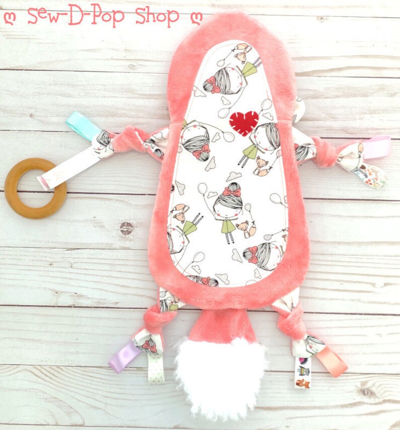 May include: A pink and white baby toy with a soft, fuzzy texture. The toy has a variety of textures and colours, including a wooden ring, ribbons, and a plush tail. The toy is decorated with a pattern of girls holding balloons.