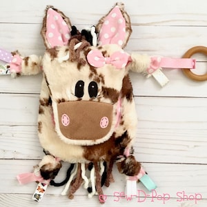 May include: A brown and white cow-print plush toy with pink ears, a pink bow, and black eyes. The toy has a brown nose and a pink mouth. The toy has a wooden ring attached to it with a pink ribbon.