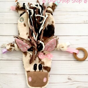 May include: A brown and white plush horse toy with pink polka dot ears and a pink bow. The horse has a wooden ring attached to it. The toy is on a white wooden background. "Sew-D-Pop Shop" is written in pink letters on the background.