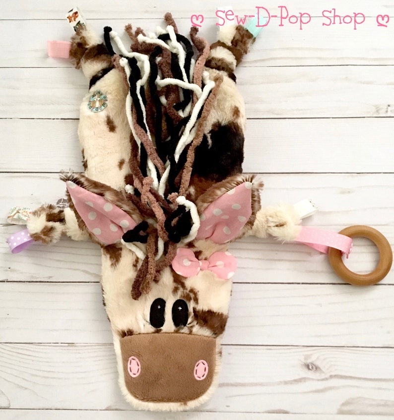 May include: A brown and white plush horse toy with pink polka dot ears and a pink bow. The horse has a wooden ring attached to it. The toy is on a white wooden background. "Sew-D-Pop Shop" is written in pink letters on the background.