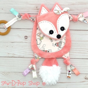 May include: A pink fox baby toy with a white and pink patterned tummy. The toy has a white fluffy tail and multiple colourful fabric loops attached.