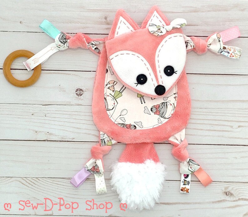 May include: A pink fox baby toy with a white and pink patterned tummy. The toy has a white fluffy tail and multiple colourful fabric loops attached.