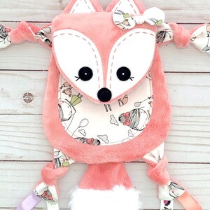 May include: A pink fox-shaped plush toy with a white face and black eyes. The toy has a white fluffy tail and is decorated with a white bow. The toy has a pocket on the front with a white background and a pattern of girls with balloons and clouds. The toy has a variety of fabric textures and is tied with ribbons.