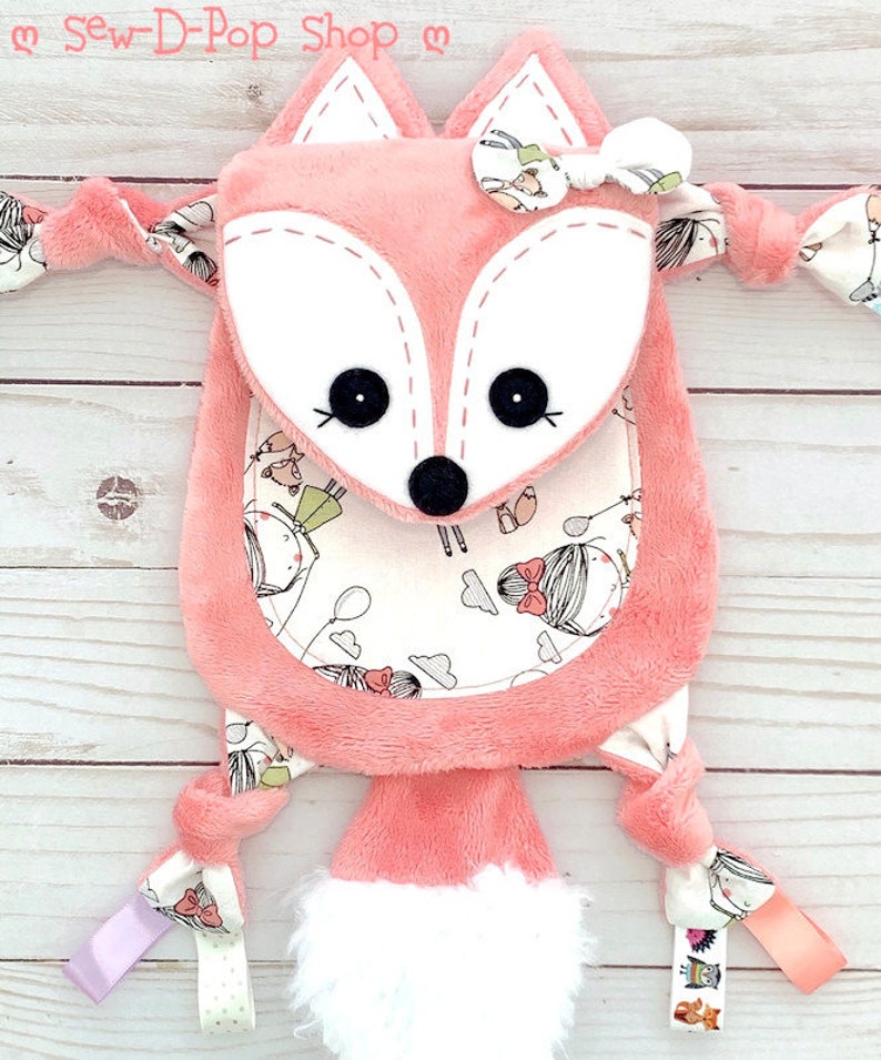 May include: A pink fox-shaped plush toy with a white face and black eyes. The toy has a white fluffy tail and is decorated with a white bow. The toy has a pocket on the front with a white background and a pattern of girls with balloons and clouds. The toy has a variety of fabric textures and is tied with ribbons.