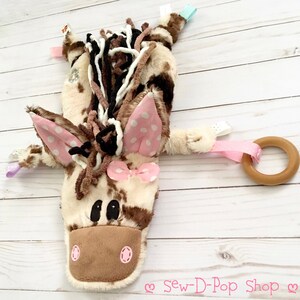 May include: A brown and white plush horse toy with pink polka dot ears and a pink bow. The toy has a wooden ring attached to it with a pink ribbon.