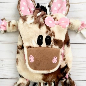 May include: A brown and white cow-print plush backpack with pink ears, a pink bow, and a pink nose. The backpack has a long tail with brown and white yarn. The backpack is on a white wooden surface. "Sew-D-Pop Shop" is written in pink letters on the top of the image.