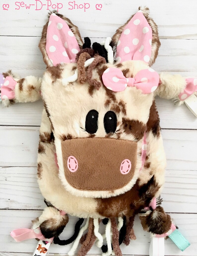 May include: A brown and white cow-print plush backpack with pink ears, a pink bow, and a pink nose. The backpack has a long tail with brown and white yarn. The backpack is on a white wooden surface. "Sew-D-Pop Shop" is written in pink letters on the top of the image.