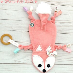 May include: A pink fox baby toy with a white tail, a wooden ring, and various fabric textures. The toy has a white face with black eyes and a black nose. The toy is on a white wooden background.