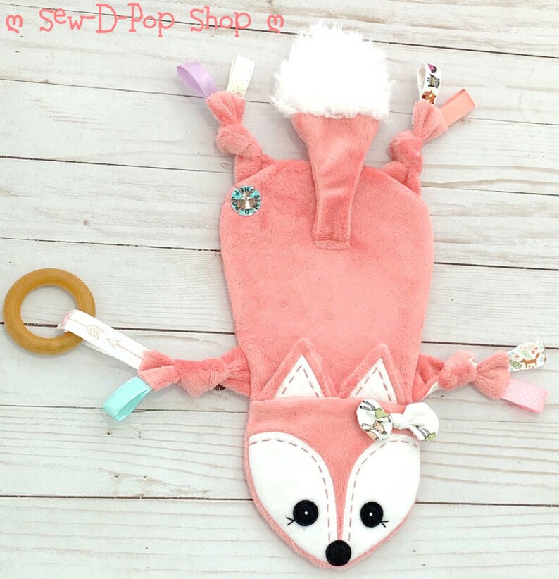 May include: A pink fox baby toy with a white tail, a wooden ring, and various fabric textures. The toy has a white face with black eyes and a black nose. The toy is on a white wooden background.