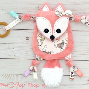 May include: A pink fox baby toy with a white face and a white fluffy tail. The toy has a variety of textures and colours, including pink, white, blue, and green. The toy also has a wooden ring and a variety of ribbons.
