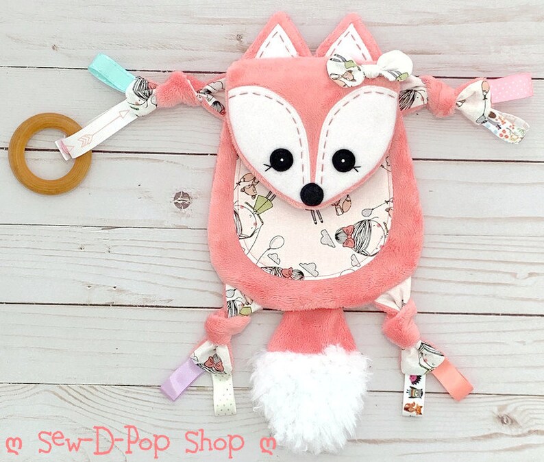 May include: A pink fox baby toy with a white face and a white fluffy tail. The toy has a variety of textures and colours, including pink, white, blue, and green. The toy also has a wooden ring and a variety of ribbons.
